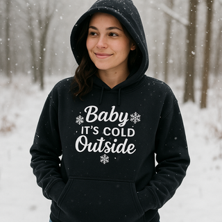 Person wearing a black hoodie with 'Baby It's Cold Outside' text in a snowy  forest. IN BLACK 