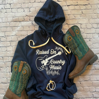 This is a Raised On Country Music Graphic Hoodie on a solid navy. The brand is Next Level and it is presented as a flat lay.