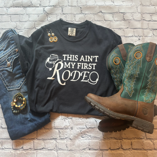 This is the This Ain't My First Rodeo Graphic Tee in the color black. The brand is Comfort Colors. It is presented as a flat lay.